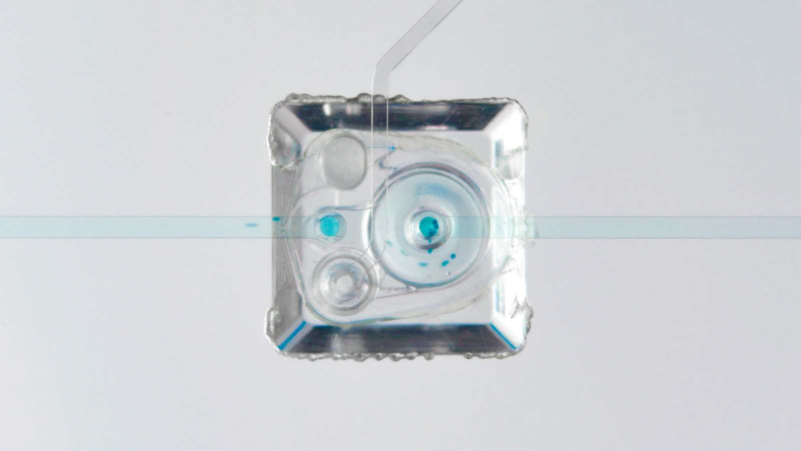 Close-up of the bottom of an MV-1 microvavle. A circular diaphragm sits within the cuboid housing. A light blue liquid with particles is flowing through the microvalve.