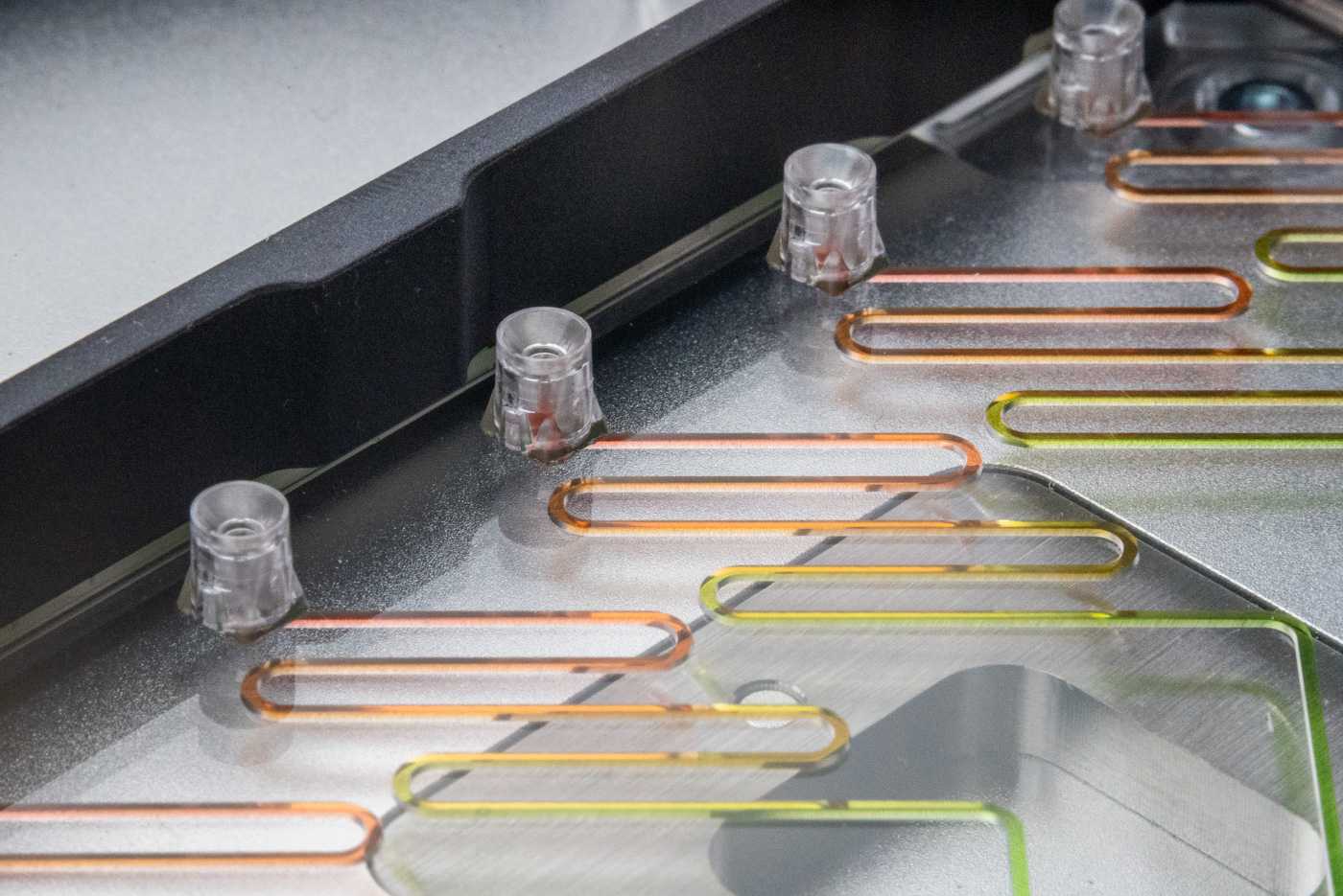 A microfluidic device with an SLAS adapter placed on the deck of an automated liquid handler. The microfluidic device has multiple pipette interfaces along one edge connected to meandering channels filled with multiple colored fluids.